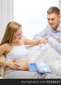 pregnancy, parenthood and celebration concept - happy family expecting child sitting on sofa with gift box and baby bootees