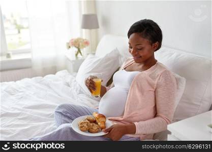 pregnancy, people and eating concept - happy pregnant african american woman drinking orange juice with pastry in bed at home. pregnant woman with orange juice and pastry in bed