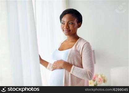pregnancy, people and expectation concept - happy pregnant african american woman looking through window at home. pregnant woman looking through window at home