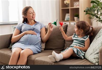 pregnancy, people and family concept - happy pregnant mother and little daughter playing with paper crane origami at home. pregnant mother and daughter with crane origami