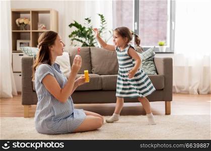 pregnancy, people and family concept - happy pregnant mother and little daughter blowing soap bubbles at home. pregnant mother and daughter blowing soap bubbles