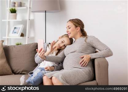 pregnancy, people and family concept - happy pregnant woman and girl sitting on sofa and taking selfie by smartphone at home. pregnant woman and girl taking smartphone selfie