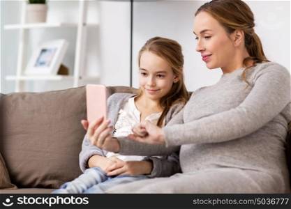 pregnancy, people and family concept - happy pregnant woman and girl sitting on sofa and taking selfie by smartphone at home. pregnant woman and girl taking smartphone selfie