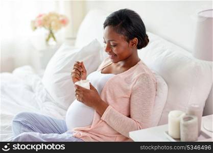 pregnancy, people and food concept - happy pregnant african american woman eating yogurt in bed at home. pregnant woman eating yogurt in bed