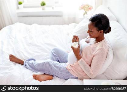 pregnancy, people and food concept - happy pregnant african american woman eating yogurt in bed at home. pregnant woman eating yogurt in bed
