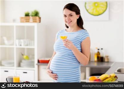 pregnancy, people and healthy eating concept - happy pregnant woman with orange slice at home kitchen. happy pregnant woman eating orange at home