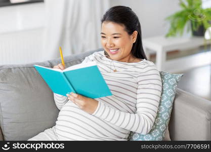 pregnancy, people and leisure concept - happy smiling pregnant asian woman sitting on sofa and writing to diary or notebook at home. happy pregnant woman writing to diary at home