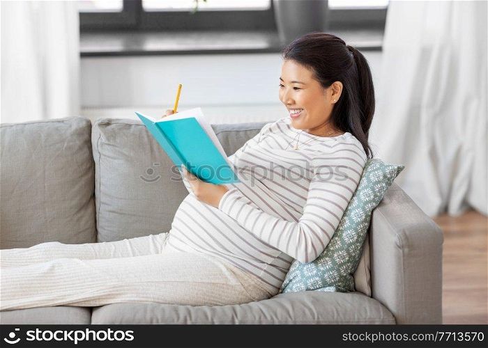pregnancy, people and leisure concept - happy smiling pregnant asian woman sitting on sofa and writing to diary or notebook at home. happy pregnant woman writing to diary at home