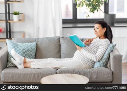 pregnancy, people and leisure concept - pregnant asian woman sitting on sofa and writing to diary or notebook at home. pregnant woman writing to diary at home