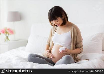 pregnancy, people and maternity concept - happy pregnant asian woman applying stretch mark cream to belly. happy asian pregnant woman applying cream to belly