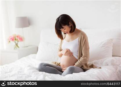 pregnancy, people and maternity concept - happy pregnant asian woman sitting in bed at home bedroom and looking at her bare belly. happy pregnant asian woman sitting in bed at home