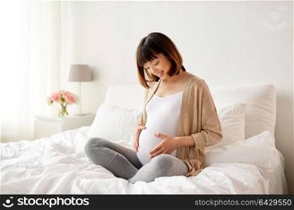 pregnancy, people and maternity concept - happy pregnant asian woman sitting in bed at home bedroom. happy pregnant asian woman sitting in bed at home
