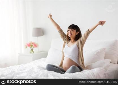 pregnancy, people and maternity concept - happy pregnant asian woman stretching in bed at home bedroom. happy pregnant asian woman sitting in bed at home