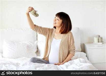 pregnancy, people and maternity concept - happy pregnant asian woman with baby bootees sitting in bed at home bedroom. happy pregnant woman with bootees in bed at home. happy pregnant woman with bootees in bed at home