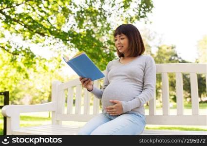 pregnancy, people and motherhood concept - happy pregnant asian woman sitting on park bench and reading book. happy pregnant asian woman reading book at park