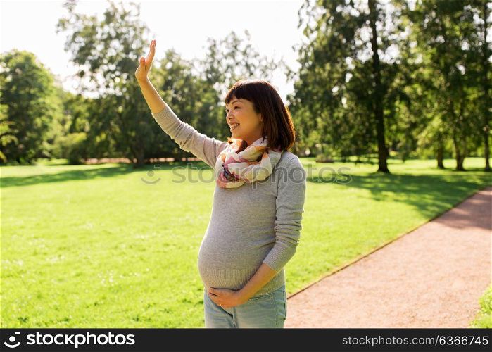 pregnancy, people and motherhood concept - happy pregnant asian woman waving hand at park. happy pregnant asian woman waving hand at park