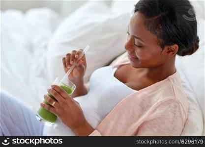 pregnancy, people and rest concept - close up of happy pregnant african american woman drinking green vegetable juice or smoothie in bed at home. pregnant woman drinking vegetable juice in bed