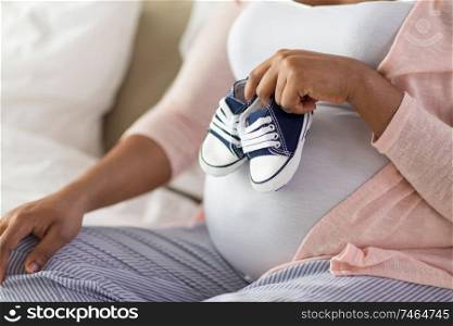 pregnancy, people and rest concept - close up of pregnant african american woman sitting on bed and holding little baby bootees at home bedroom. pregnant african woman with little baby bootees