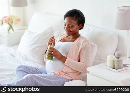 pregnancy, people and rest concept - happy pregnant african american woman drinking green vegetable juice or smoothie in bed at home. pregnant woman drinking vegetable juice in bed