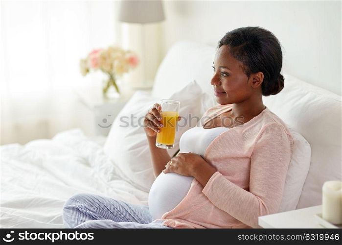 pregnancy, people and rest concept - happy pregnant african american woman drinking orange juice in bed at home. pregnant woman drinking orange juice in bed
