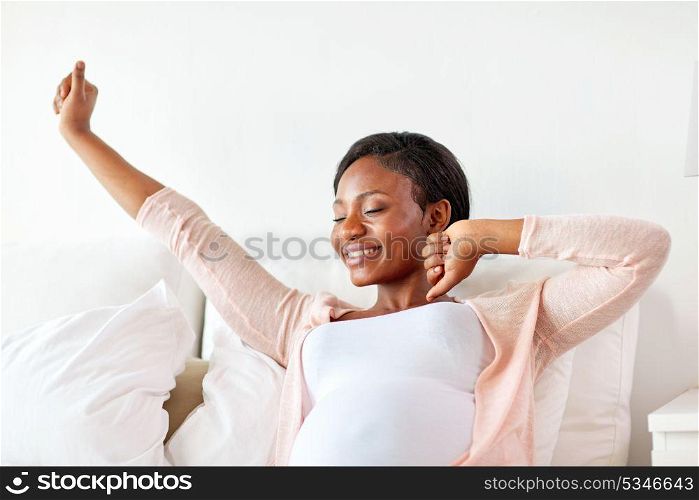 pregnancy, people and rest concept - happy pregnant african american woman stretching in bed at home. happy pregnant african woman stretching in bed