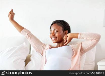 pregnancy, people and rest concept - happy pregnant african american woman stretching in bed at home. happy pregnant african woman stretching in bed
