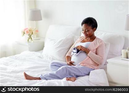 pregnancy, people and rest concept - happy pregnant african american woman with baby bootees in bed at home bedroom. pregnant african woman with baby bootees in bed