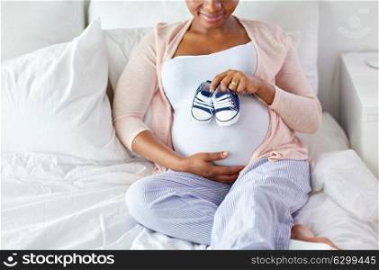 pregnancy, people and rest concept - happy pregnant african american woman with baby bootees in bed at home bedroom. pregnant african woman with baby bootees in bed