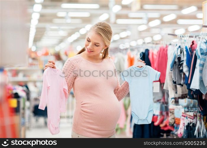 pregnancy, people and shopping concept - happy pregnant woman choosing between blue and pink baby bodysuits at clothing store. pregnant woman with baby clothes at clothing store
