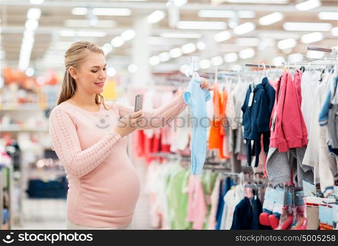 pregnancy, people and shopping concept - happy pregnant woman taking picture of blue baby bodysuit with smartphone at clothing store. pregnant woman taking picture of baby clothes
