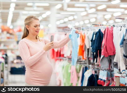 pregnancy, people and shopping concept - happy pregnant woman taking picture of blue baby bodysuit with smartphone at clothing store. pregnant woman taking picture of baby clothes