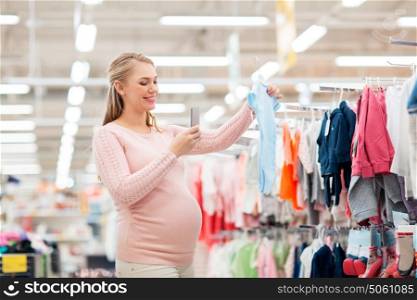 pregnancy, people and shopping concept - happy pregnant woman taking picture of blue baby bodysuit with smartphone at clothing store. pregnant woman taking picture of baby clothes