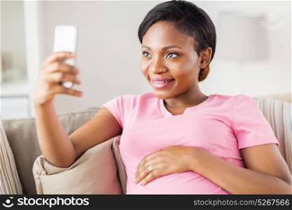 pregnancy, people and technology concept - happy pregnant african american woman with smartphone taking selfie at home. pregnant woman taking smartphone selfie at home