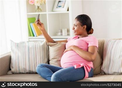 pregnancy, people and technology concept - happy pregnant african american woman with smartphone taking selfie at home. pregnant woman taking smartphone selfie at home