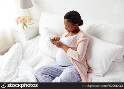 pregnancy, people and technology concept - happy pregnant african american woman with smartphone in bed at home. happy pregnant woman with smartphone at home
