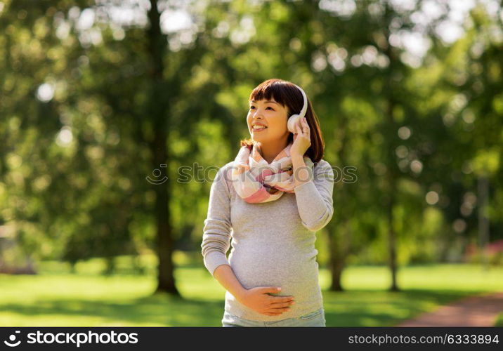 pregnancy, people and technology concept - happy pregnant asian woman in headphones at park listening to music. happy pregnant asian woman in headphones at park