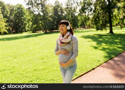 pregnancy, people and technology concept - happy pregnant asian woman in headphones walking at park and listening to music. happy pregnant asian woman in headphones at park