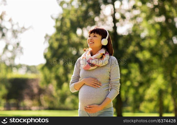 pregnancy, people and technology concept - happy pregnant asian woman in headphones at park listening to music. happy pregnant asian woman in headphones at park
