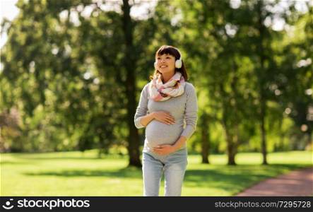 pregnancy, people and technology concept - happy pregnant asian woman in headphones walking at park and listening to music. happy pregnant asian woman in headphones at park