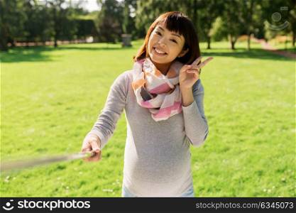 pregnancy, people and technology concept - happy pregnant asian woman with selfie stick taking picture at park. happy pregnant asian woman taking selfie at park