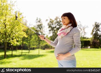 pregnancy, people and technology concept - happy pregnant asian woman with smartphone selfie stick taking picture at park. happy pregnant asian woman taking selfie at park