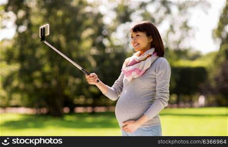 pregnancy, people and technology concept - happy pregnant asian woman with smartphone selfie stick taking picture at park. happy pregnant asian woman taking selfie at park