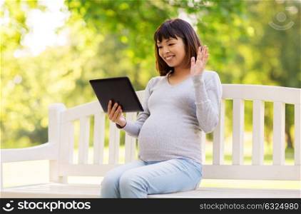 pregnancy, people and technology concept - happy pregnant asian woman with tablet pc computer sitting on park bench and having video chat. happy pregnant asian woman with tablet pc at park