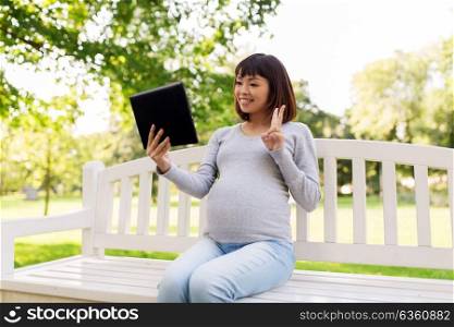 pregnancy, people and technology concept - happy pregnant asian woman with tablet pc computer sitting on park bench and having video chat. happy pregnant asian woman with tablet pc at park