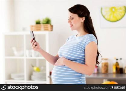 pregnancy, people and technology concept - happy pregnant woman with smartphone taking selfie at home kitchen. happy pregnant woman with smartphone at kitchen