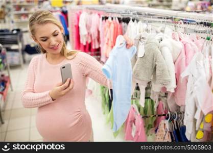 pregnancy, people, technology and shopping concept - happy pregnant woman taking picture of blue baby bodysuit with smartphone at clothing store. pregnant woman taking picture of baby clothes