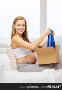 pregnancy, post, delivery and motherhood concept - smiling pregnant woman sitting on sofa and opening parcel box with blue cardigan