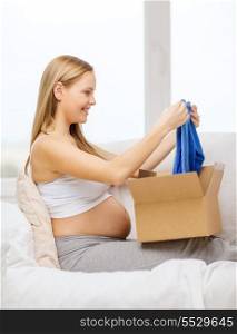 pregnancy, post, delivery and motherhood concept - smiling pregnant woman sitting on sofa and opening parcel box with blue cardigan