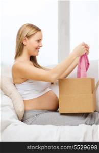 pregnancy, post, delivery and motherhood concept - smiling pregnant woman sitting on sofa and opening parcel box with pink cardigan