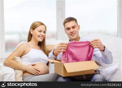 pregnancy, post, delivery and parenthood concept - happy family expecting child sitting on sofa and opening parcel box with pink cardigan